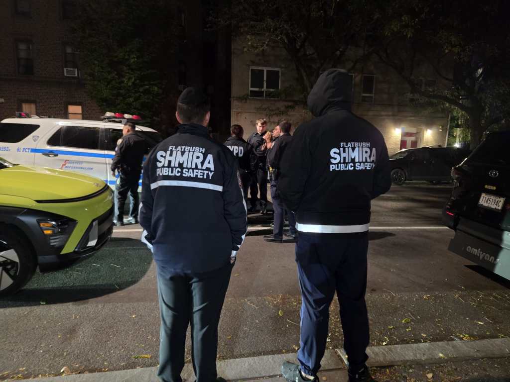 Two Flatbush Shmira Public Safety members observing police officers on a street at night