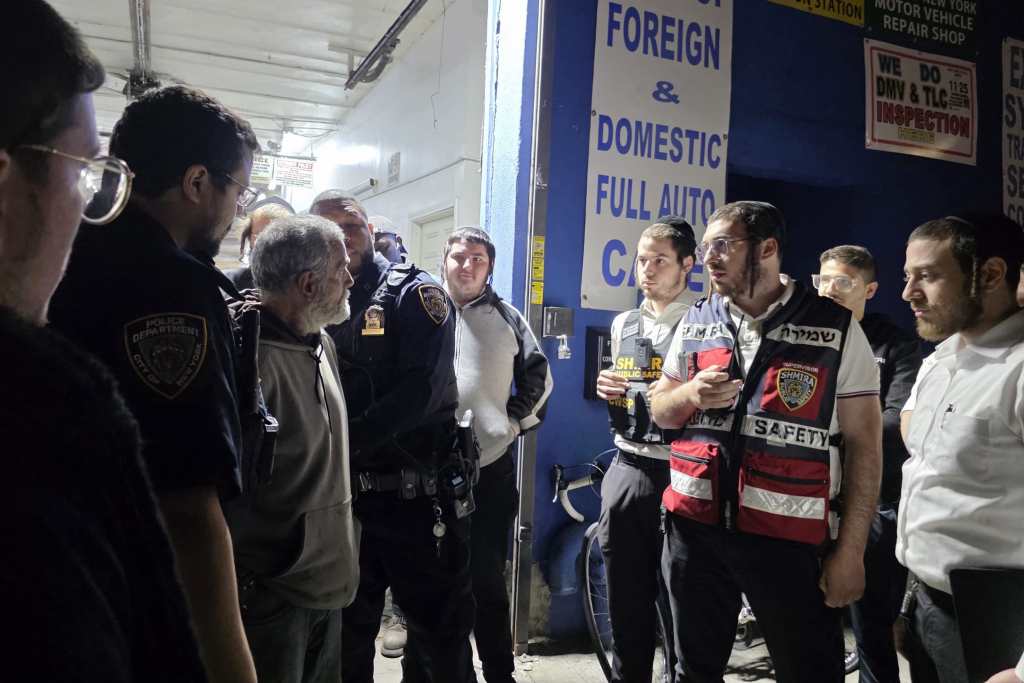 Police officers and Shmira safety members standing around a man near an auto repair shop.
